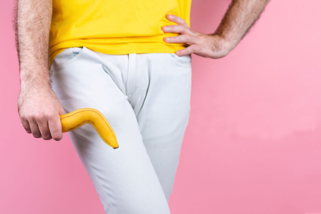 Impotence. A man in white jeans is holding a broken banana near his genitals with his legs together and his hand on his waist.